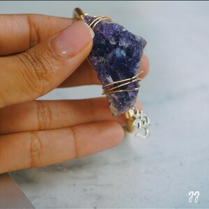 May include: A gold wire wrapped amethyst crystal pendant with a silver Om charm. The pendant is held in a hand.
