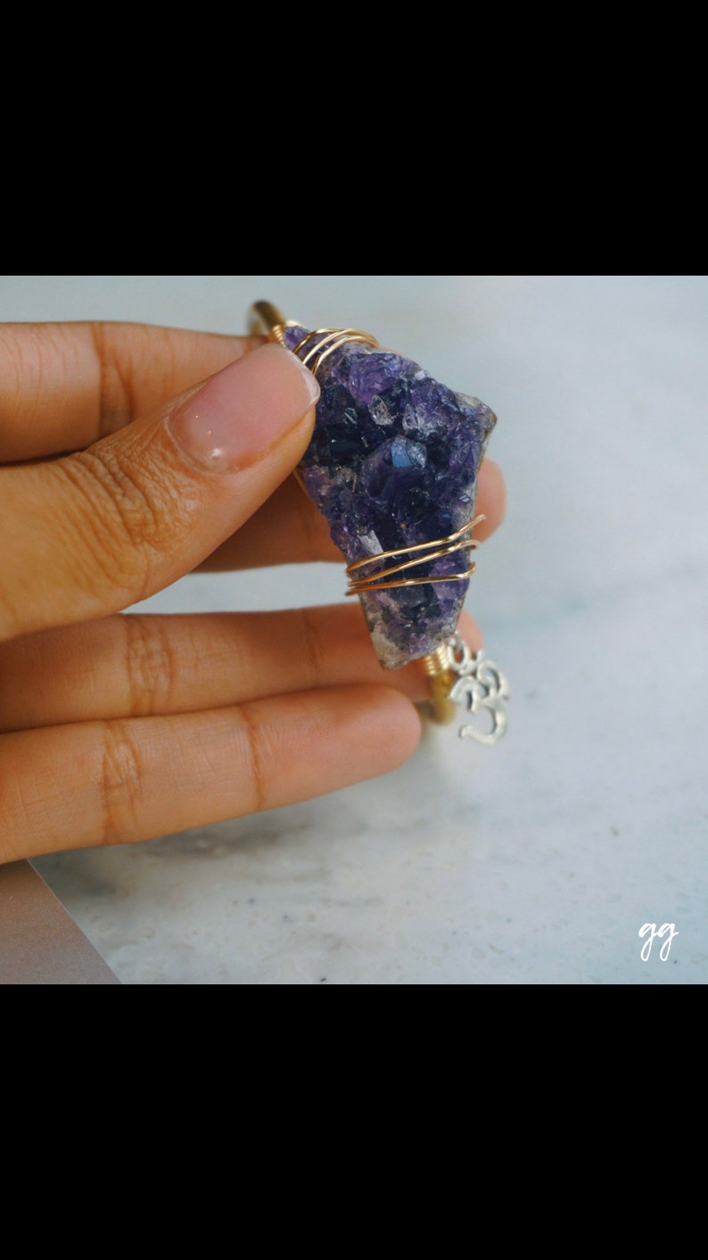 May include: A gold wire wrapped amethyst crystal pendant with a silver Om charm. The pendant is held in a hand.