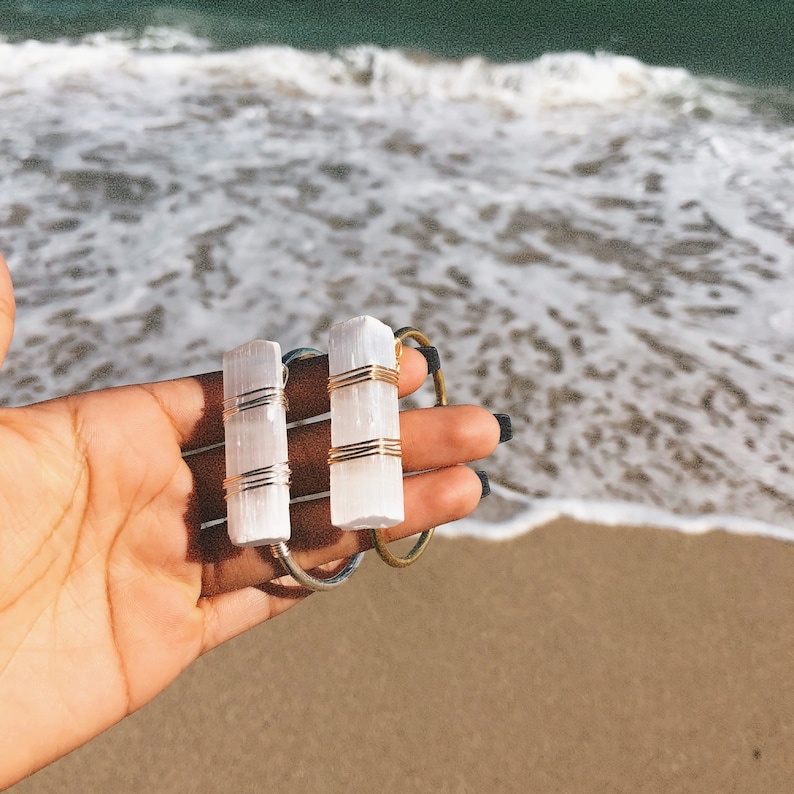 May include: Three white selenite crystal bracelets wrapped in gold wire. The bracelets are stacked on top of each other and are being held by a hand.
