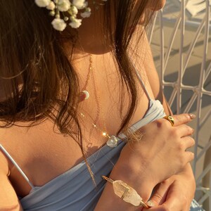 May include: A woman wearing a light blue top with a gold necklace and a crystal pendant. She is also wearing a gold ring and a crystal bracelet.