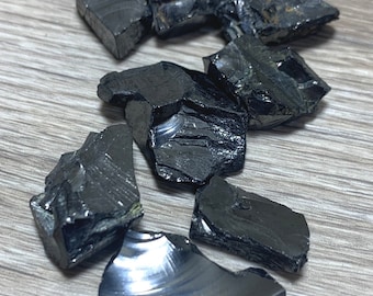 Könnte beinhalten: Eine Ansammlung unregelmäßig geformter, dunkler Obsidiansteine mit glänzender, reflektierender Oberfläche. Die Steine variieren in der Größe und sind auf einer hellen Holzmaserung verstreut, was einen Kontrast erzeugt.