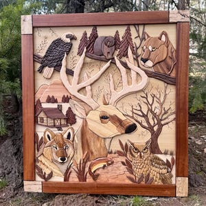 May include: Wooden wall art featuring a detailed relief carving of wildlife. The artwork includes a deer, eagle, bear, cougar, wolf, owl, and a fish, set against a backdrop of trees, mountains, and a cabin. The frame is a dark brown color.