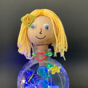 Puede incluir: Una botella de vidrio con una cabeza de muñeca de madera y cabello de lana amarilla. La botella contiene canicas azules, una flor amarilla, un amuleto de rayo y luces de cadena.