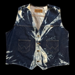 May include: A dark blue denim vest with a tie-dye pattern. The vest has a V-neck, two front pockets, and a button-up closure. The Wrangler logo is visible on the chest.