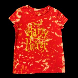 May include: A short-sleeved crew neck t-shirt featuring a red and white tie-dye pattern. The front displays the gold-colored text Harry Potter in a stylized font, accompanied by a small gold owl graphic and a wand icon above the lettering.
