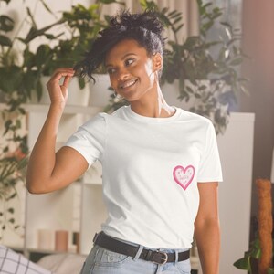 May include: White t-shirt with a pink heart graphic on the chest. The heart contains the text "Sweet as" and small star details. The shirt is paired with blue jeans and a black belt.