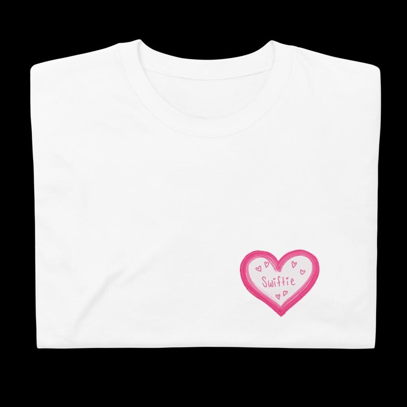 May include: White t-shirt with a pink heart design. The heart is outlined in a darker pink and contains the word "Swiftie" in the center, along with small hearts. The shirt is folded.