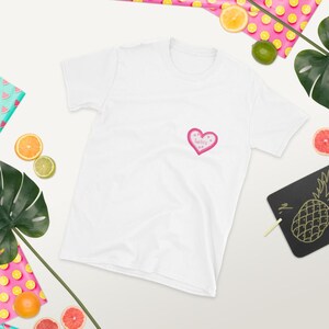 May include: White t-shirt with a pink heart-shaped graphic on the chest. The heart has a pink outline and contains the words "Be Still" in a handwritten style. The shirt is surrounded by tropical fruit and leaves.