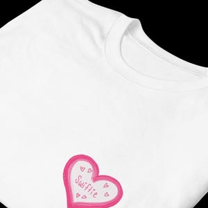 May include: White t-shirt featuring a pink heart design with the word "Swiftie" in cursive. The heart is outlined in pink and has small heart details. The shirt is made of a soft material.