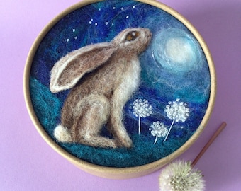 May include: A needle felted artwork of a brown hare looking up at a white moon in a blue night sky. The hare is surrounded by white dandelion seeds.
