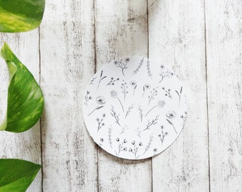 May include: A white round coaster with a black and white floral pattern. The coaster is on a white wooden surface with a green plant in the background.