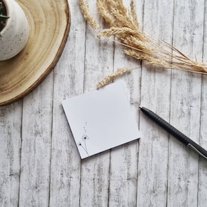 Può includere: Un blocco note bianco con un semplice disegno di dente di leone nero, una penna nera e un mazzo di spighe di grano secche su uno sfondo di legno sbiancato.