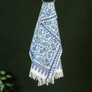 May include: A white cotton throw blanket with a blue paisley pattern and fringe trim. The blanket is folded and hanging from a hook.