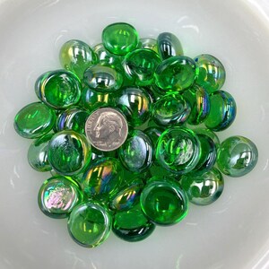 May include: A collection of green glass marbles with a coin for size comparison, arranged in a white bowl. The marbles have a glossy finish and reflect light, creating a shimmering effect. The marbles are approximately 2 cm in diameter.