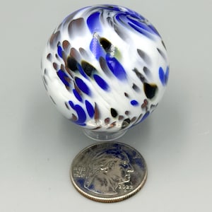 Puede incluir: Una canica de vidrio esférica con una base blanca y patrones en espiral de color azul cobalto, negro y marrón. La canica está colocada directamente sobre una moneda estadounidense de 25 centavos, que sirve como referencia de escala.