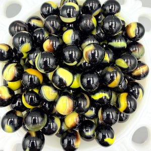 May include: A collection of glossy black marbles with swirls of yellow, brown, and cream colors. The marbles are displayed in a white, scalloped-edge bowl, creating a vintage aesthetic. These marbles are perfect for collectors or as decorative items.