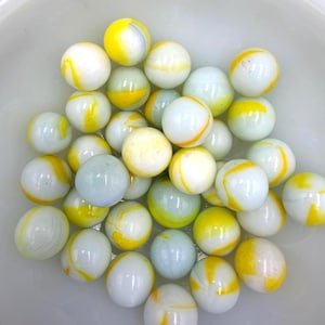 Puede incluir: Una colección de canicas de vidrio esféricas con una base blanca translúcida y patrones de remolinos en amarillo brillante y azul pálido. Las canicas están agrupadas, mostrando superficies lisas y reflectantes con colores variados.