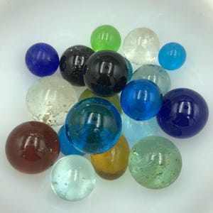 May include: A collection of glass marbles in various colors, including blue, green, clear, black, and brown. The marbles are of different sizes and have a glossy finish, reflecting light. The marbles are scattered on a white surface.