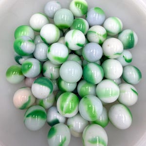 Puede incluir: Un conjunto de canicas de vidrio blanco y verde arremolinado en un cuenco blanco. Las canicas tienen un acabado brillante y diferentes tonos de verde y blanco. Un juguete clásico.