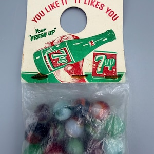 May include: Vintage 7 Up soda bottle and can graphic on a cardboard tag with the text "You like it, it likes you." A clear plastic bag contains a collection of colorful marbles. The tag has a hole for hanging.