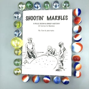 May include: A book titled "Shootin' Marbles: A Rule Book & Brief History, 22 Games for Marbles" by Sue & Jack Hahn, surrounded by a variety of marbles, including clear, green, yellow, and red, white, and blue marbles.