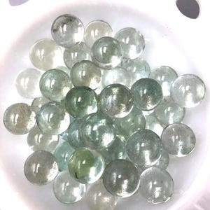 Può includere: Una collezione di numerose piccole biglie di vetro sferiche con una tonalità chiara e verde pallido. Le biglie sono raggruppate, mostrando superfici lisce e riflettenti con bolle d'aria interne, riposte all'interno di una ciotola bianca con fori circolari.