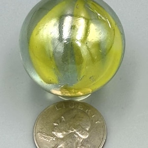 May include: A clear glass marble with internal yellow swirls positioned above a 1985 United States quarter coin, which serves as a scale reference for the size of the spherical object.