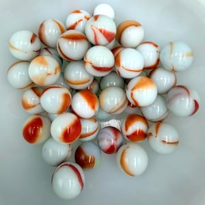 May include: A collection of white marbles with vibrant red and orange swirls, filling a white bowl. The marbles vary in their patterns, creating a dynamic visual texture. The marbles are smooth and spherical, reflecting light.