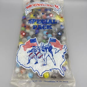 Puede incluir: Una bolsa de plástico transparente llena de canicas de vidrio de colores variados. El empaque presenta el texto Bicentennial 1776-1976 Special Pack, una ilustración del mapa de Estados Unidos, dos banderas estadounidenses y tres figuras tocando tambores.