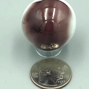 May include: A glossy, maroon-colored sphere with a white band, resting on a clear stand. A U.S. quarter coin is next to the sphere for size comparison. The coin shows the words "In God We Trust" and the year 2022.