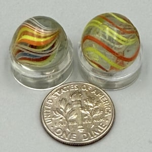May include: Two clear glass marbles with internal swirling patterns of yellow, orange, and white, each resting on a small circular clear stand. A United States one dime coin is placed in front of the marbles for a size reference.