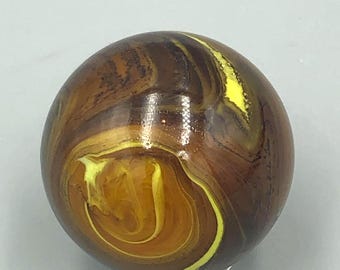 May include: A brown and yellow swirled glass marble. The marble has a glossy finish and a pattern of swirling brown and yellow colors. The marble is a sphere and sits on a silver metal ring.