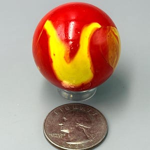 May include: A spherical glass marble with a vibrant red base color and swirling yellow patterns throughout its surface. The marble is positioned directly above a United States quarter coin, which serves as a scale reference for its size.