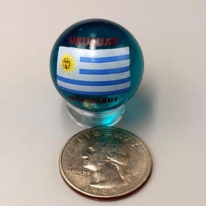 May include: A translucent teal glass marble featuring the flag of Uruguay with a yellow sun and blue and white stripes. The word URUGUAY is printed in red above the flag and MARVELOUS in red below. A 1985 US quarter is placed next to the marble.