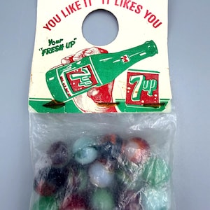 May include: A vintage 7-Up branded cardboard header card with the text "YOU LIKE IT IT LIKES YOU" and "Your FRESH UP" featuring a green soda bottle illustration, attached to a clear plastic bag containing a collection of assorted colorful glass marbles.