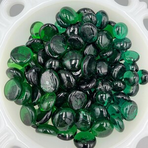May include: A white, scalloped bowl is filled with a collection of glossy, emerald green glass pebbles. The smooth, rounded stones vary in shade, creating depth. These decorative pebbles are ideal for crafts and home decor.