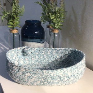 May include: A blue and white patterned rope basket with a decorative knot on the side. The basket is sitting on a white surface with three glass vases and green plants in the background.
