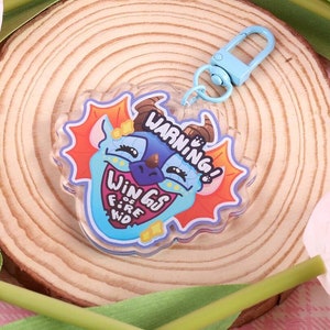 May include: A blue and orange cartoon dragon with the text "WARNING! WINGS OF FIRE KID" on a clear acrylic keychain with a blue metal ring.