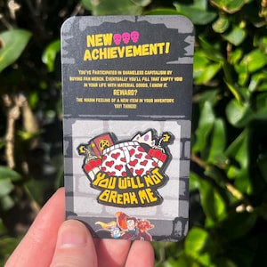 May include: A novelty pin featuring a cartoon design with the text "YOU WILL NOT BREAK ME." The pin is on a card with the text "NEW ACHIEVEMENT!" and a humorous description about buying merchandise. The pin has a red, white, and yellow color scheme.