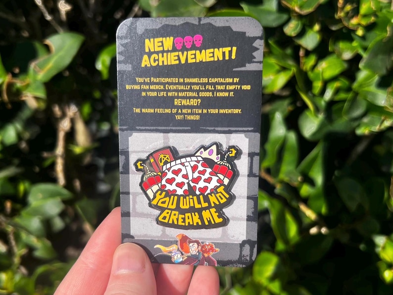 May include: A novelty pin featuring a cartoon design with the text "YOU WILL NOT BREAK ME." The pin is on a card with the text "NEW ACHIEVEMENT!" and a humorous description about buying merchandise. The pin has a red, white, and yellow color scheme.