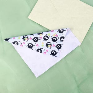May include: A white, triangular cloth with a repeating pattern of black cartoon characters with white eyes and pink flowers. A light yellow envelope rests on a light green surface.