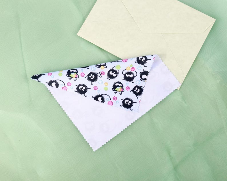 May include: A white, triangular cloth with a repeating pattern of black cartoon characters with white eyes and pink flowers. A light yellow envelope rests on a light green surface.