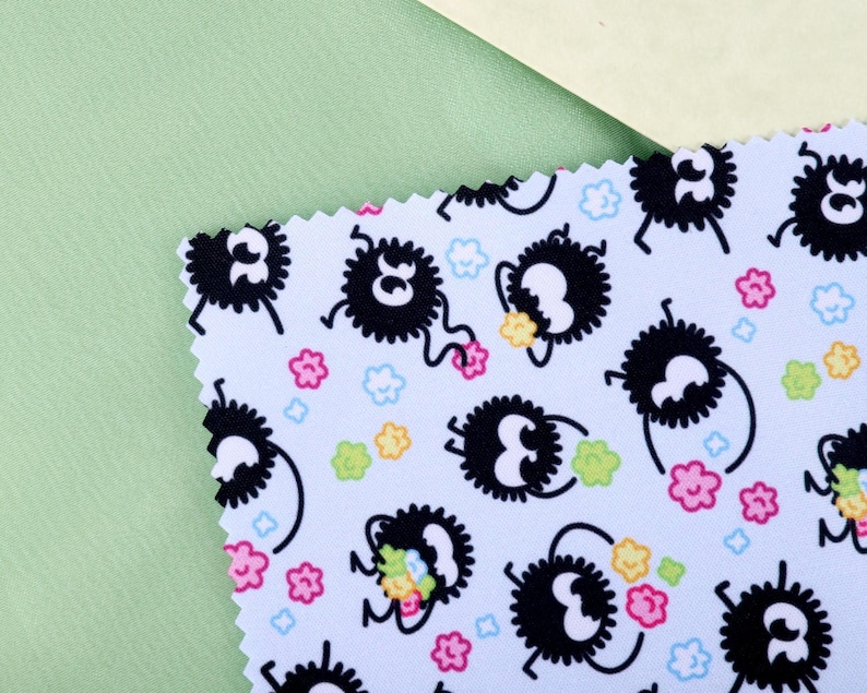 May include: A light blue fabric sample with a repeating pattern of black, fluffy creatures with white eyes and legs, holding colorful flowers. The design includes pink, yellow, and blue floral accents. The fabric is set against a light green and cream background.