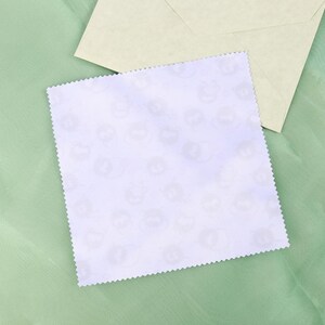 May include: A square, white card with a scalloped edge, featuring a repeating pattern of light gray cat faces. An off-white envelope rests on a light green fabric background.