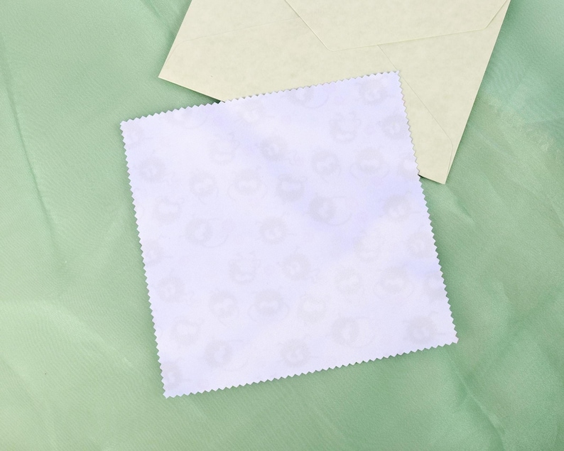 May include: A square, white card with a scalloped edge, featuring a repeating pattern of light gray cat faces. An off-white envelope rests on a light green fabric background.