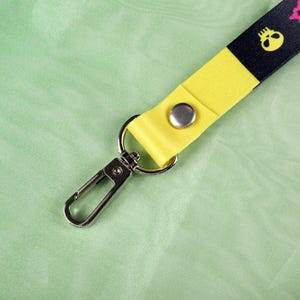 May include: A lanyard with a black and yellow design featuring a skull pattern. The lanyard has a yellow strap with a silver snap and a metal clasp. The background is a light green fabric.