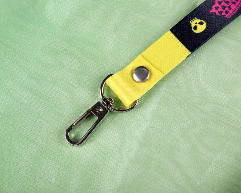 May include: A lanyard with a black and yellow design featuring a skull pattern. The lanyard has a yellow strap with a silver snap and a metal clasp. The background is a light green fabric.