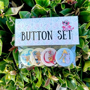 May include: A set of four round buttons with colorful fantasy character designs, packaged in a clear plastic bag. The packaging features the text "BUTTON SET" and a pink dragon illustration. The background is lush green foliage.