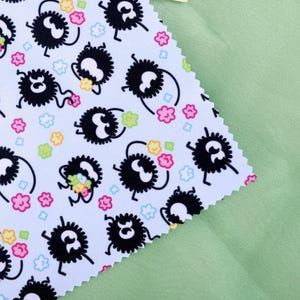 May include: A light blue fabric with a repeating pattern of black, fluffy creatures with white eyes and legs, holding colorful flowers. Pink, yellow, and green flowers and small blue clouds are scattered across the fabric. A light green fabric is in the background.