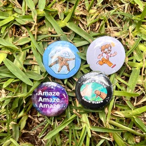 Puede incluir: Cuatro chapas circulares con diseños distintos: una azul con una figura en pose, una blanca con un personaje corriendo, una morada con temática de galaxia y el texto Amaze Amaze Amaze, y una negra con una ilustración de manos.